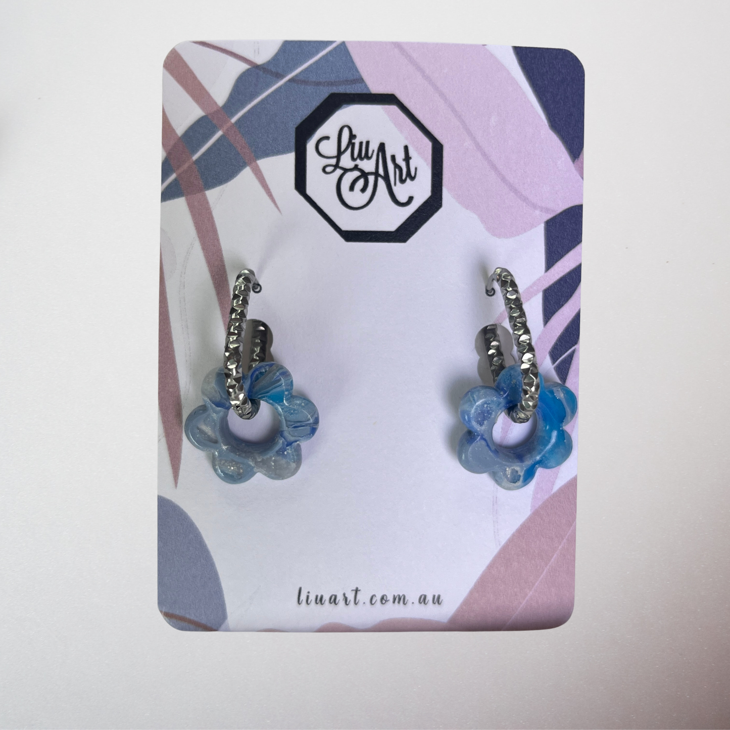 A pair of silver huggie hoop earrings with blue flower-shaped beads, displayed on a branded 'Liu Art' card against a plain light background.