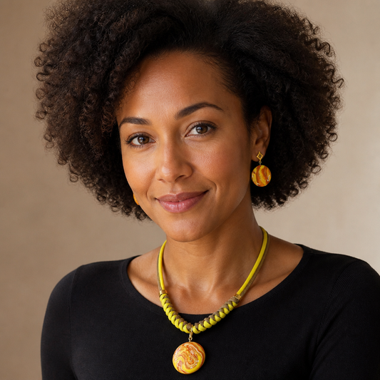 A woman with curly dark hair wearing a yellow beaded necklace with a round pendant and matching yellow earrings, photographed against a plain beige studio background.