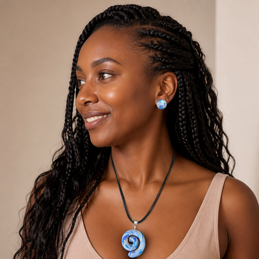 A woman with long black braided hair is shown in a close-up portrait, wearing a black cord necklace with a blue spiral pendant and a matching blue earring against a plain beige background.
