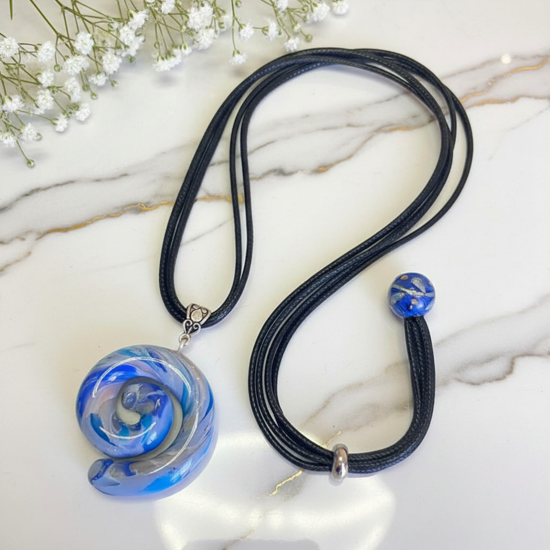 A black multi-strand cord necklace with a large blue and white marbled spiral pendant, displayed on a white marble surface with small white flowers in the corner.