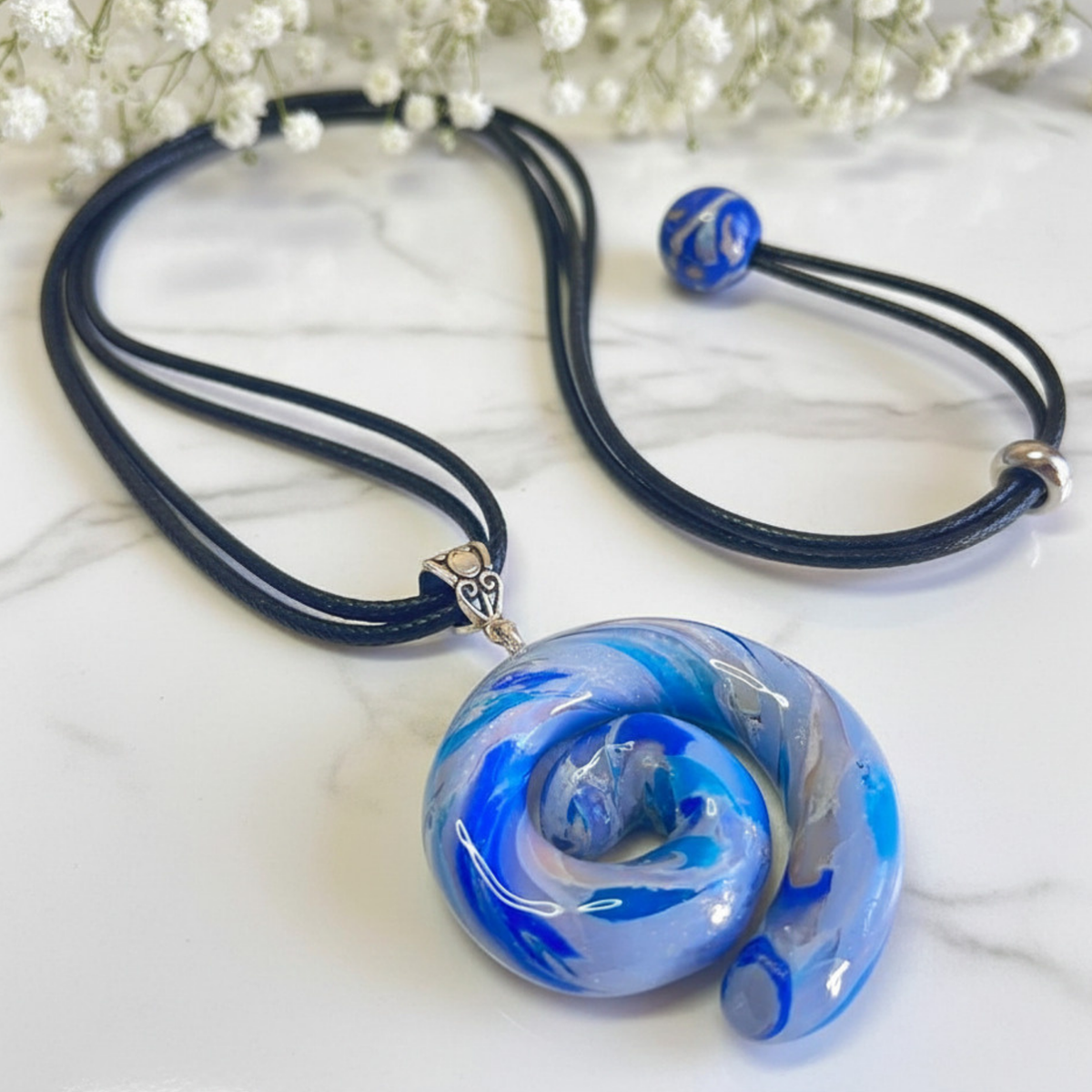 A handmade glass pendant necklace featuring a swirling blue, white, and light gray glass charm on a black cord, displayed on a white marble surface with baby's breath flowers in the background.