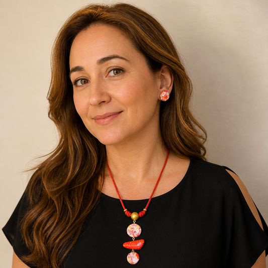A woman with long wavy brown hair wearing a red cord necklace with pink and orange shell-like beads and matching pink earrings, against a plain light beige background.