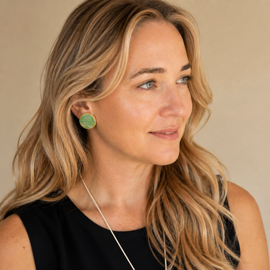 A woman with blonde hair wearing a black sleeveless top and a delicate silver necklace, featuring a green circular stud earring on one ear. She is shown in profile against a solid beige background.