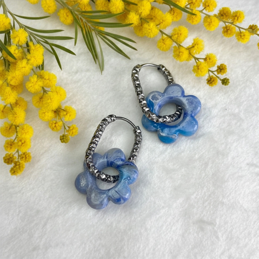 A pair of dangling hoop earrings with blue flower-shaped charms and silver-toned beaded hoops, arranged on a white textured surface with yellow flowers in the corner.
