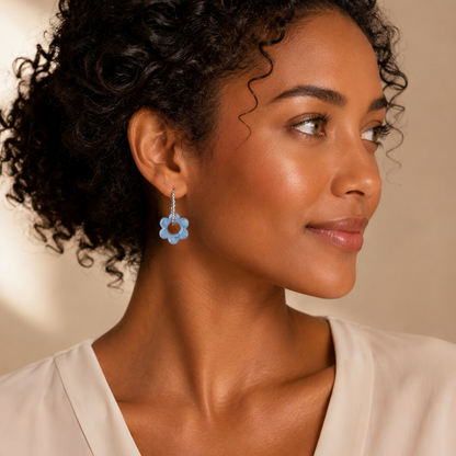 A close-up of a woman with curly dark hair wearing a light blue flower-shaped dangle earring, dressed in a cream-colored top against a warm beige background.