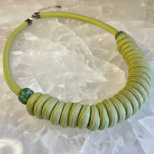 A handmade coiled cord necklace in a light olive green color, featuring a textured spiral design and a decorative green bead accent, displayed on a white textured stone background.