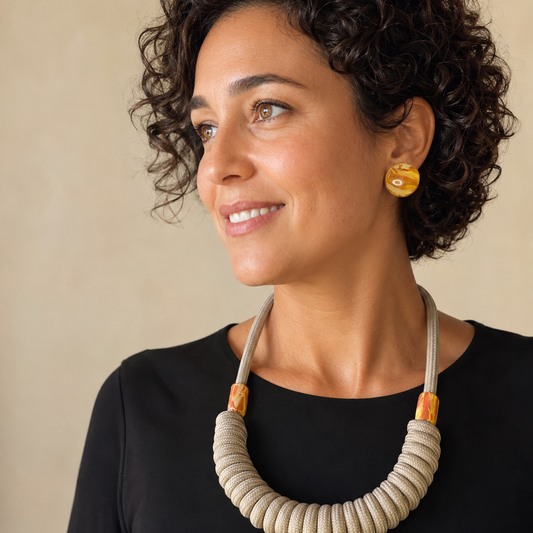 A woman with short curly dark hair is shown in a close-up portrait, wearing a chunky coiled beige statement necklace and a small round yellow earring against a plain beige background.