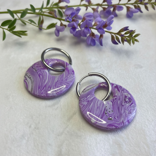 Two circular purple and white marbled resin earrings with silver-tone split rings, displayed on a white textured surface with purple flowers in the background.