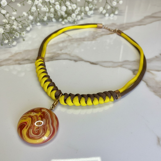 A handmade corduroy necklace featuring yellow and brown woven cords, a gold-tone clasp, and a round marbled pendant with red, yellow, and white swirls, displayed on a white marble surface with baby's breath flowers.