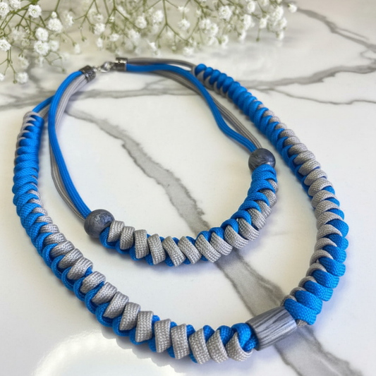 A two-tone blue and gray paracord necklace displayed on a white marble surface with small white flowers in the background.