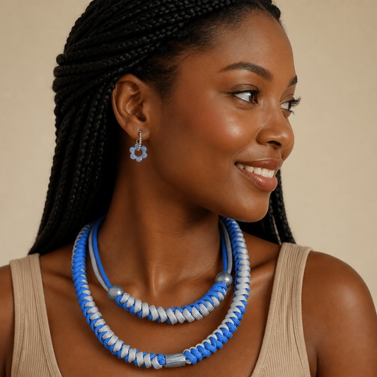 A woman with black braided hair is shown in profile wearing a blue and white beaded necklace and matching flower-shaped earring against a warm beige background.