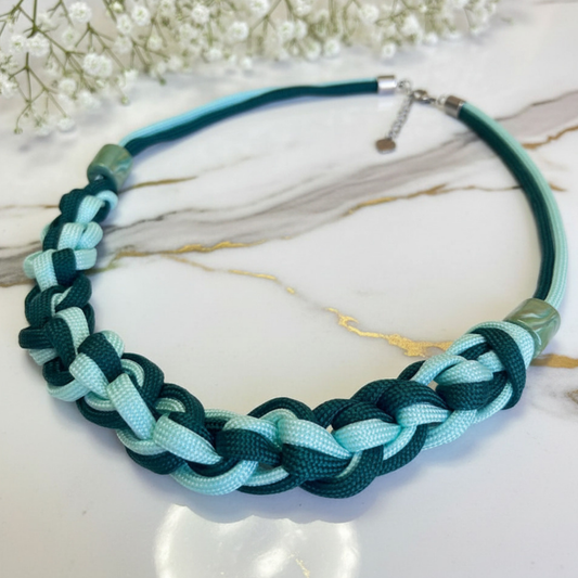 A braided cord choker necklace in dark teal and light aqua, displayed on a white marble surface with small white flowers in the background.