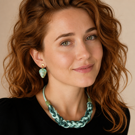 A close-up portrait of a woman with wavy red hair wearing a chunky braided mint green and white necklace and a matching teardrop-shaped drop earring with gold-toned metal.