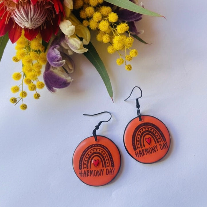 Orange earrings with 'Harmony Day' text on a white background with flowers.