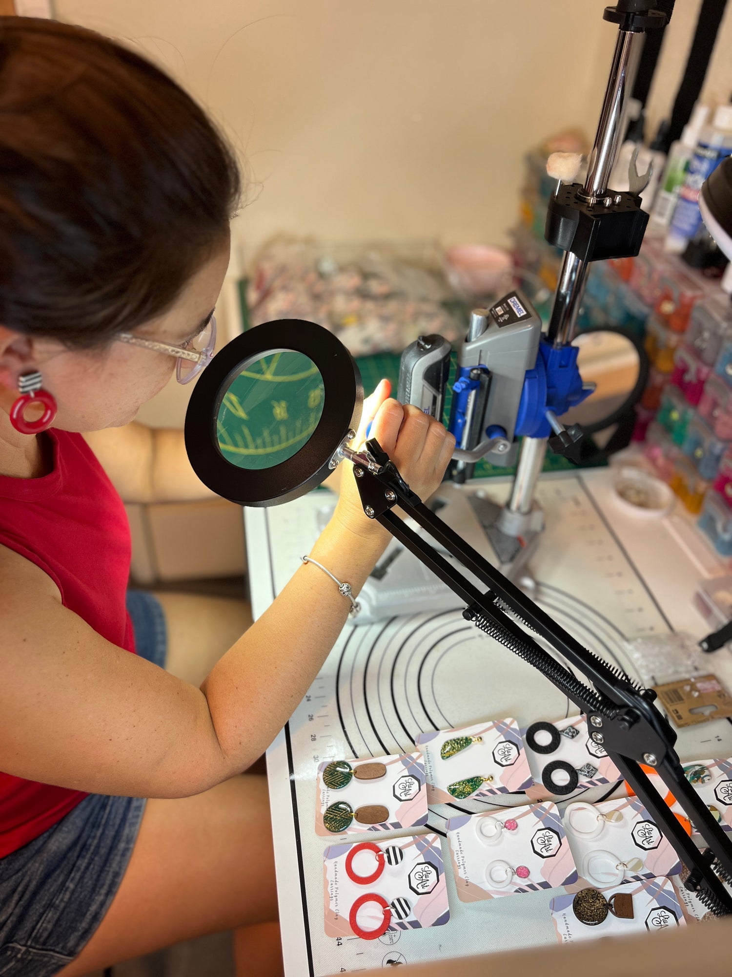 A person wearing glasses and a red top uses a magnifying loupe and clamp to examine small items on a work surface in a cluttered craft or workshop environment.